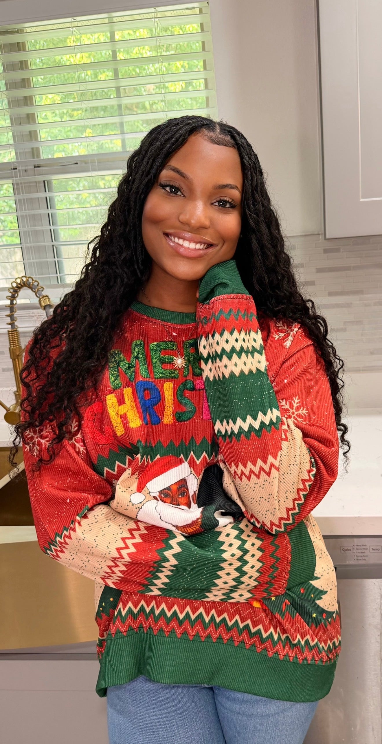 A woman wearing a festive Christmas sweater with 'MERRY CHRIST' in colorful letters, standing in a kitchen setting.